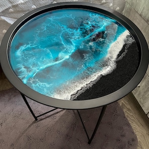 Op de afbeelding: Ronde bijzettafel met een zwart metalen frame en een hars top. Het tafelblad heeft een blauw en wit golfpatroon, met een zwarte rand die op een strand lijkt. De tafel heeft drie zwarte metalen poten.