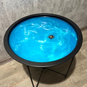 May include: A round side table with a black metal frame and a blue resin top with white swirls. A small turtle figurine sits in the centre of the table top.