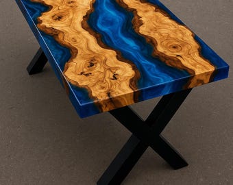 Puede incluir: Mesa de centro rectangular con un diseño único. La encimera presenta un río de resina azul que fluye entre dos secciones de madera marrón claro. La mesa está sostenida por patas de metal negro cruzadas.
