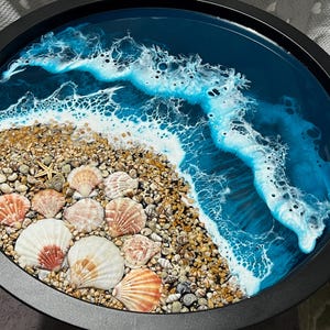 May include: Circular black tray with a coastal design. The tray depicts a beach scene with seashells, sand, and a blue ocean with white foamy waves. A starfish is also visible on the beach. The tray is a decorative item.