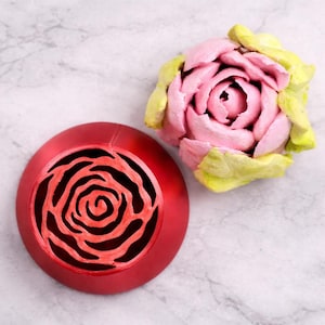 Könnte beinhalten: Ein rotes, kreisförmiges Dekorationsobjekt mit einem rosenförmigen Ausschnitt neben einem rosa-gelben rosenförmigen Ornament. Die Objekte befinden sich auf einer weiß-grau marmorierten Oberfläche. Das Rosenornament hat rosa Blütenblätter und gelbe Blätter.