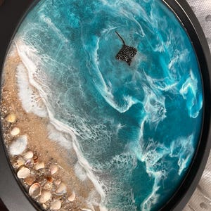 May include: Circular resin artwork showing a beach scene. It features a sandy shore with seashells, leading to turquoise ocean waves. A black and white spotted stingray swims in the water. The piece is framed in a black circular border.
