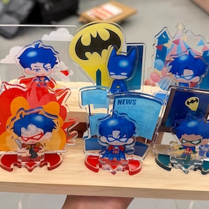 May include: A collection of six acrylic character standees featuring chibi-style figures with blue hair, displayed on a light wood tiered stand. The figures include various superhero-themed backgrounds, a yellow bat symbol, and a blue newspaper graphic.
