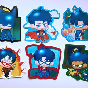 May include: A set of six die-cut stickers featuring chibi-style comic book characters. The designs include Batman with a yellow bat signal, Superman with a white dog, Nightwing with balloons, Red Hood in an explosion, Robin, and a character in a library.