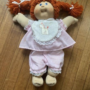 May include: A vintage doll with red yarn pigtails tied with white ribbons. The doll has blue eyes, a smiling expression, and is wearing a pink gingham outfit with a white bib featuring a giraffe applique. The doll's outfit includes a dress and matching pants.