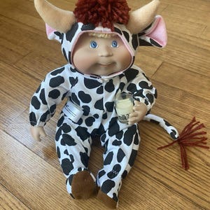 May include: A Cabbage Patch Kid doll dressed in a black and white cow costume with a red pom-pom on its head. The doll is holding a small glass with the word "milk?" printed on it. The doll has blue eyes and is sitting on a wooden surface.