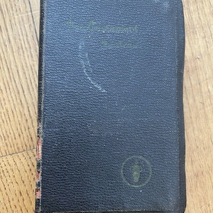 May include: A small, dark blue book with a textured cover. The book has a green emblem and text on the cover. The edges of the book show wear and tear. The book is resting on a wooden surface.