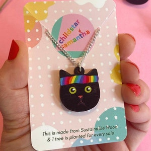 May include: A black wooden cat pendant necklace with a rainbow headband. The necklace is on a silver chain and is displayed on a card with the text "childstar samantha" and "This is made from Sustainable Wood & 1 tree is planted for every sale".