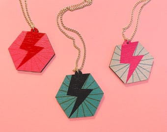 May include: Three necklaces with hexagonal pendants featuring lightning bolt designs. One pendant is red and pink, one is black and teal, and one is pink and silver.