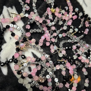 May include: A collection of elastic bracelets featuring pink, black, and translucent beads interspersed with silver-toned letter beads spelling out various words and phrases, accented with small metallic cross charms throughout the design.