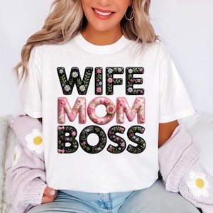 Op de afbeelding: Wit t-shirt met de tekst "WIFE MOM BOSS" in een bloemenontwerp. Het woord "WIFE" is in zwart met witte bloemen, "MOM" is in roze met roze bloemen en "BOSS" is in zwart met roze bloemen.