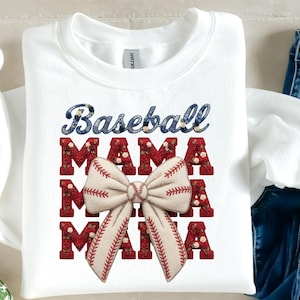Puede incluir: Sudadera blanca con las palabras "Baseball MAMA" en un diseño decorativo. La palabra "Baseball" es azul con detalles florales, y "MAMA" es roja con detalles florales. Un lazo con temática de béisbol está centrado debajo del texto.