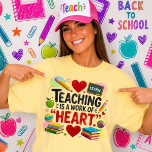 May include: A pale yellow t-shirt featuring a graphic with the text "TEACHING IS A WORK OF HEART" alongside illustrations of a pencil, a red heart, a chalkboard, and stacks of books. A pink and white trucker hat with the word "Teach" is also visible.