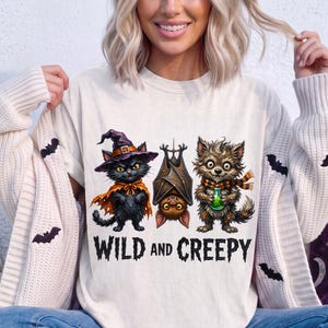 May include: A cream-colored t-shirt featuring a graphic of a black cat in a purple witch hat, a brown bat hanging upside down, and a furry creature holding a green potion bottle, with the text WILD AND CREEPY printed in black below the characters.
