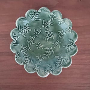 May include: A green ceramic dish with a scalloped edge, adorned with numerous embossed sea turtle designs. The dish has a glossy finish and is set against a brown wooden surface. The turtles vary in size and are evenly distributed across the surface.