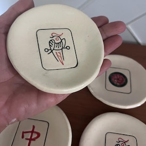 May include: A set of round, off-white ceramic dishes featuring hand-painted designs inspired by mahjong tiles. One dish displays a black and red bird illustration, another shows a red Chinese character, and a third features a red floral pattern.
