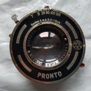 Op de afbeelding: Een zwarte en zilveren camera-sluiter met de tekst "PRONTO" en "Gauthier 0. m. b. H. Calmbach" erop gedrukt. De sluiter heeft een ronde opening met een glazen lens en een wijzerplaat met nummers van 25 tot 125.