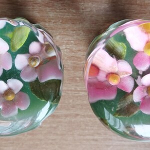 Può includere: Due fermacarte in vetro con un motivo floreale. Ogni fermacarte è rotondo e presenta fiori rosa e bianchi con centri gialli, su uno sfondo verde. I fermacarte hanno un diametro di circa 7,6 cm.