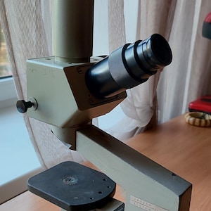 May include: A vintage microscope with a gray and black body. The microscope has a black stage and a silver knob on the side. The microscope is sitting on a wooden table.