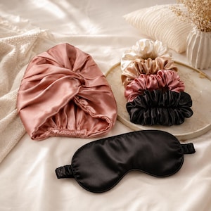 Op de afbeelding: Een oudroze satijnen slaapmuts met een geplooide voorkant, een zwart satijnen slaapmasker met een elastische band, en een stapel van vier satijnen haarelastiekjes in crème, champagne, oudroze en zwart, allemaal met een gladde afwerking.