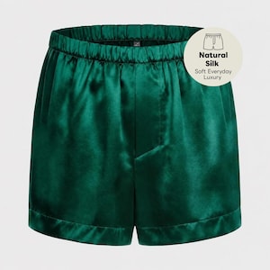 May include: Emerald green silk shorts with an elasticized waistband and a smooth, lustrous finish. A circular badge in the upper right corner features an icon of shorts and the text: Natural Silk, Soft Everyday Luxury.