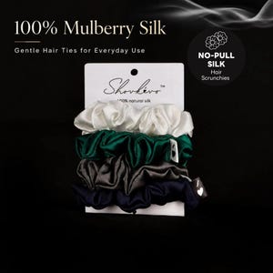 Könnte beinhalten: Ein Set aus vier Haargummis in Weiß, Smaragdgrün, Anthrazit und Marineblau, präsentiert auf einer weißen Karte mit der Aufschrift Shovkevo 100% natural silk. Die Verpackung enthält den Text 100% Mulberry Silk und No-Pull Silk Hair Scrunchies.