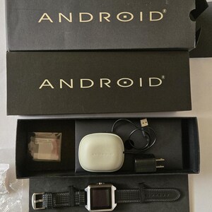 May include: An Android smartwatch and accessories are displayed in an open black box. The box contains a smartwatch with a black leather band, a charging case, a charging cable, and a wall adapter. The word "ANDROID" is printed on the box.