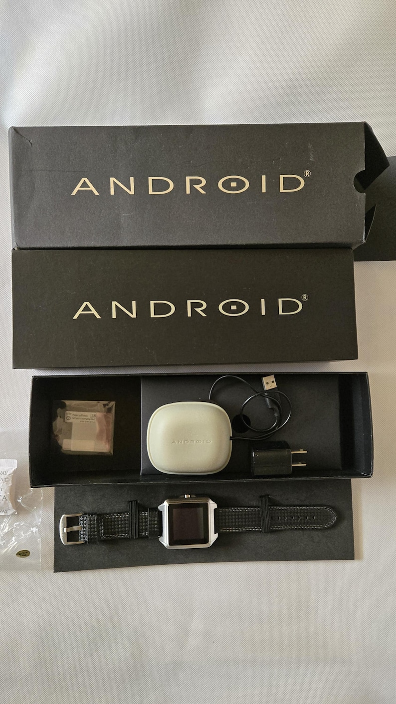 May include: An Android smartwatch and accessories are displayed in an open black box. The box contains a smartwatch with a black leather band, a charging case, a charging cable, and a wall adapter. The word "ANDROID" is printed on the box.