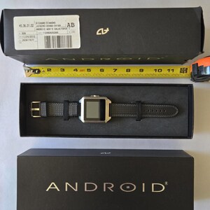 May include: A black box with the word "ANDROID" in gold lettering. Inside, a black smartwatch with a silver face and black band is displayed. A measuring tape shows the watch is approximately 10 inches long.