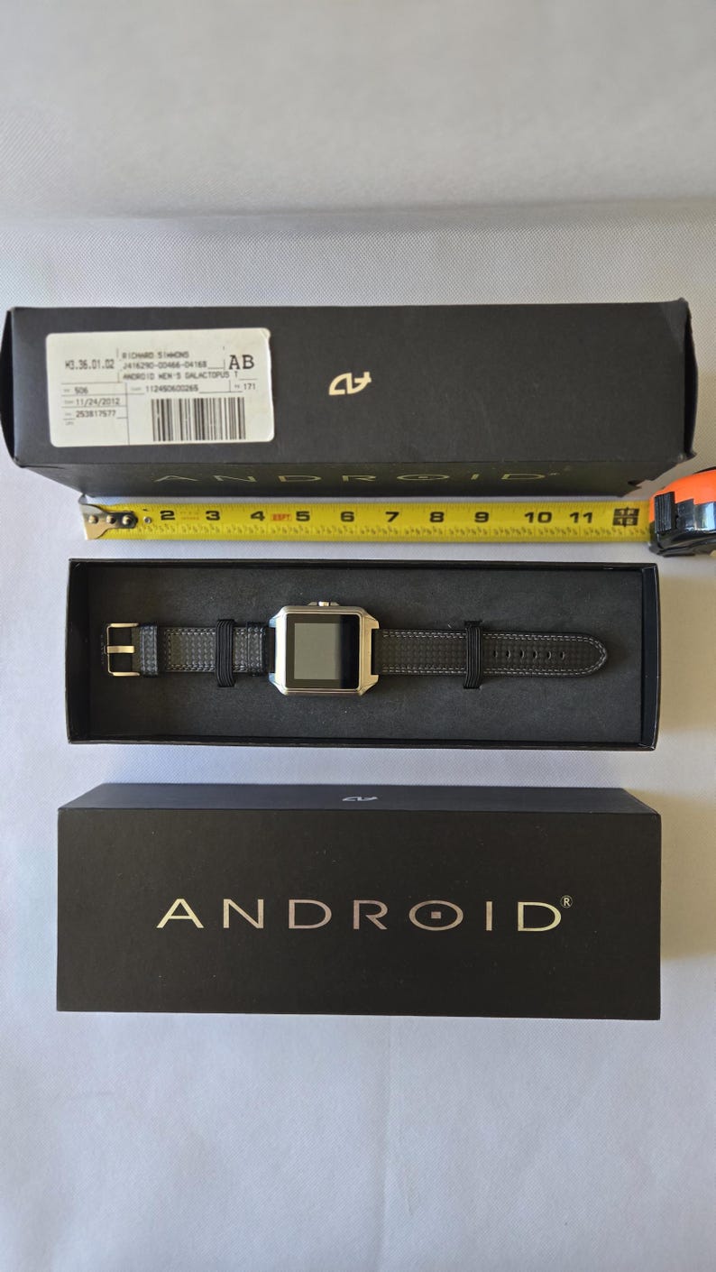May include: A black box with the word "ANDROID" in gold lettering. Inside, a black smartwatch with a silver face and black band is displayed. A measuring tape shows the watch is approximately 10 inches long.