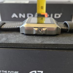 May include: A silver smartwatch with a square face and a dark gray band. The watch is resting on a black surface. The watch has a button with the letters "AD" on it. The box behind the watch has the word "ANDROID" printed on it.