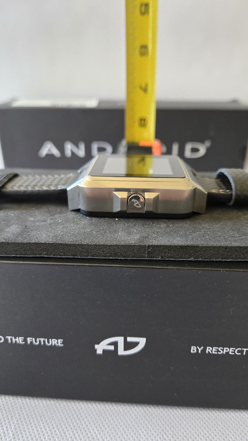May include: A silver smartwatch with a square face and a dark gray band. The watch is resting on a black surface. The watch has a button with the letters "AD" on it. The box behind the watch has the word "ANDROID" printed on it.