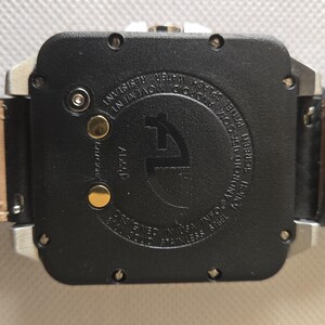 May include: Close-up of a black watch with a leather strap. The watch face is square with a silver frame. The back of the watch has text and a logo. The watch strap is brown leather with the words "GENUINE LEATHER" printed on it.