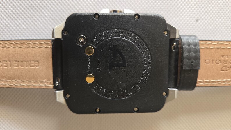 May include: Close-up of a black watch with a leather strap. The watch face is square with a silver frame. The back of the watch has text and a logo. The watch strap is brown leather with the words "GENUINE LEATHER" printed on it.