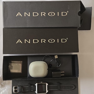 May include: An open black box set containing an Android smartwatch and accessories. The watch has a square silver face and a black leather band. Included are a charging cable, a charging adapter, and a white charging case.