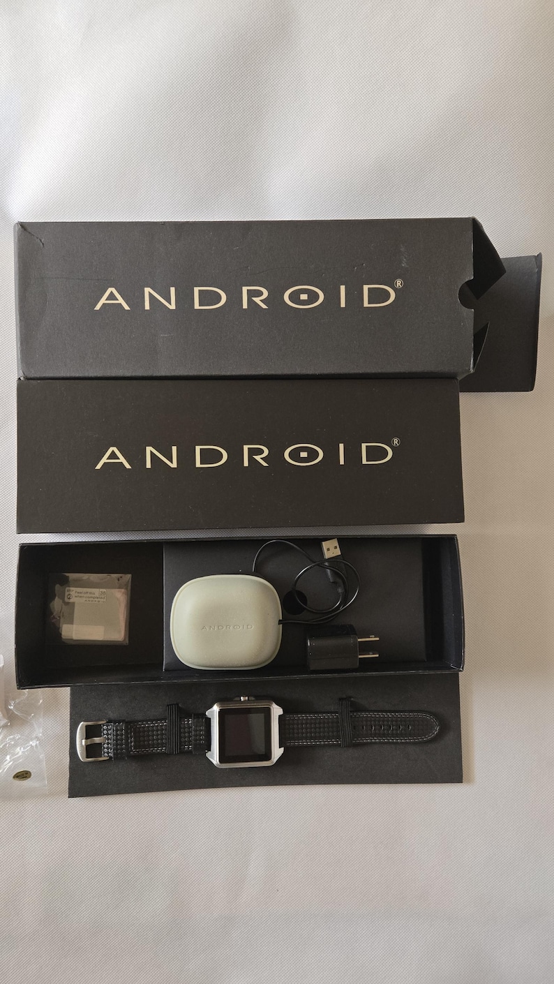 May include: An open black box set containing an Android smartwatch and accessories. The watch has a square silver face and a black leather band. Included are a charging cable, a charging adapter, and a white charging case.