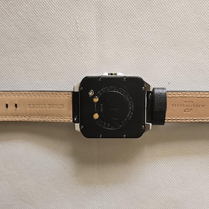 May include: Black square smartwatch with a black leather band and a silver clasp. The watch face is black with two gold-colored contact points. The underside of the band is tan with the words "GENUINE LEATHER" and "ANDROID" printed on it.