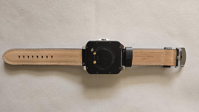 May include: Black square smartwatch with a black leather band and a silver clasp. The watch face is black with two gold-colored contact points. The underside of the band is tan with the words "GENUINE LEATHER" and "ANDROID" printed on it.