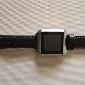 May include: A silver smartwatch with a black leather band. The watch has a square face with a dark screen and a buckle closure. The band is stitched with white thread.