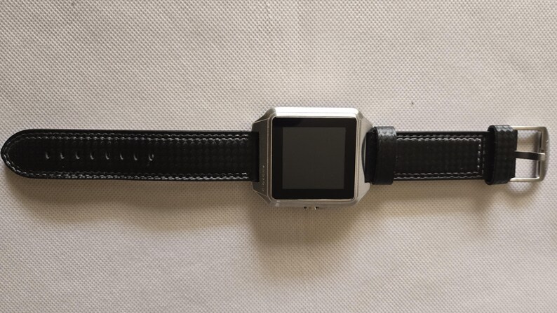 May include: A silver smartwatch with a black leather band. The watch has a square face with a dark screen and a buckle closure. The band is stitched with white thread.