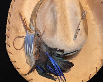 May include: A tan cowboy hat with a decorative feather and ribbon arrangement. The hat features a brown stitched brim and a silver pin. The feathers are blue, black, and brown.