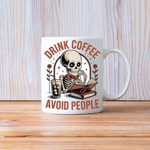 Puede incluir: Taza de cer&aacute;mica blanca con una ilustraci&oacute;n de un esqueleto leyendo un libro y bebiendo caf&eacute; helado. La taza tiene el texto "DRINK COFFEE AVOID PEOPLE" en marr&oacute;n. La taza est&aacute; sobre una superficie de madera.