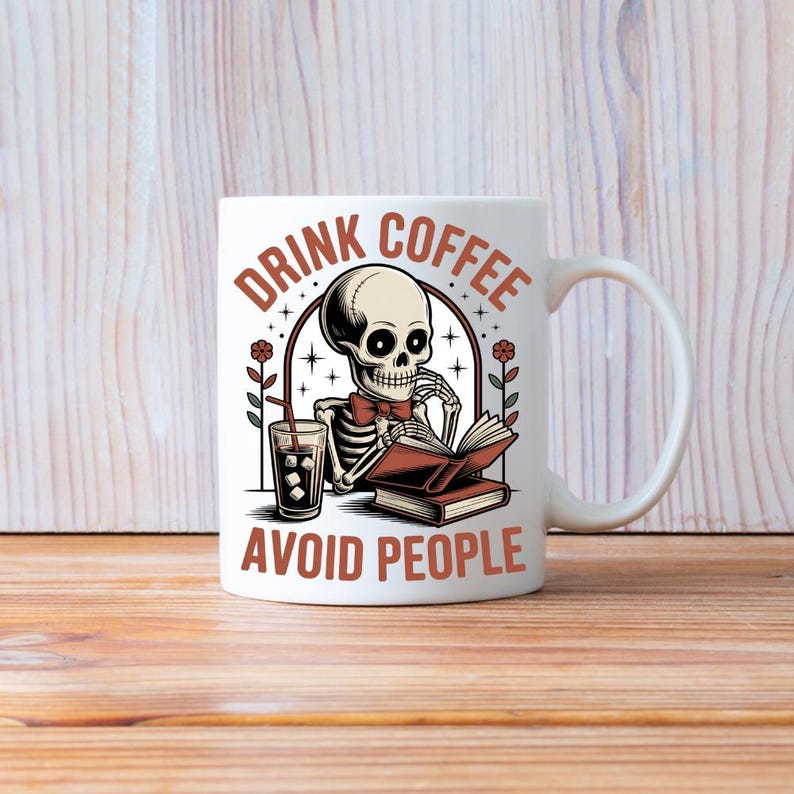 Puede incluir: Taza de cer&aacute;mica blanca con una ilustraci&oacute;n de un esqueleto leyendo un libro y bebiendo caf&eacute; helado. La taza tiene el texto "DRINK COFFEE AVOID PEOPLE" en marr&oacute;n. La taza est&aacute; sobre una superficie de madera.