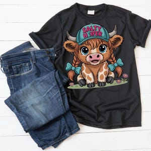 May include: Black t-shirt with a cartoon cow graphic wearing a blue hat that says "Salty Heifer". The cow has pigtails with blue bows. The shirt is paired with blue denim jeans.