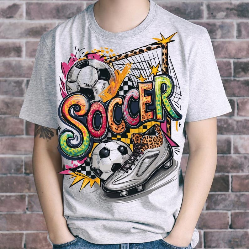 May include: A light gray t-shirt featuring a colorful soccer-themed graphic. The design includes soccer balls, a soccer cleat with a leopard print, and the word "SOCCER" in vibrant, graffiti-style lettering. The shirt is short-sleeved.