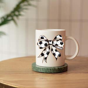 May include: White ceramic mug featuring a black and white soccer ball bow design. The mug has a curved handle and sits on a green coaster. The mug is on a wooden table.