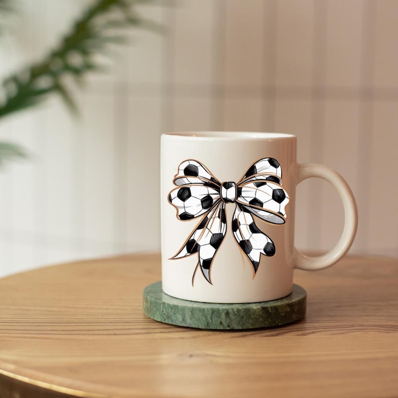 May include: White ceramic mug featuring a black and white soccer ball bow design. The mug has a curved handle and sits on a green coaster. The mug is on a wooden table.