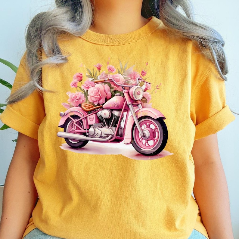 May include: Yellow t-shirt featuring a pink motorcycle design adorned with pink flowers. The motorcycle has a classic design with a round headlight and a detailed engine. The flowers add a feminine touch to the motorcycle.