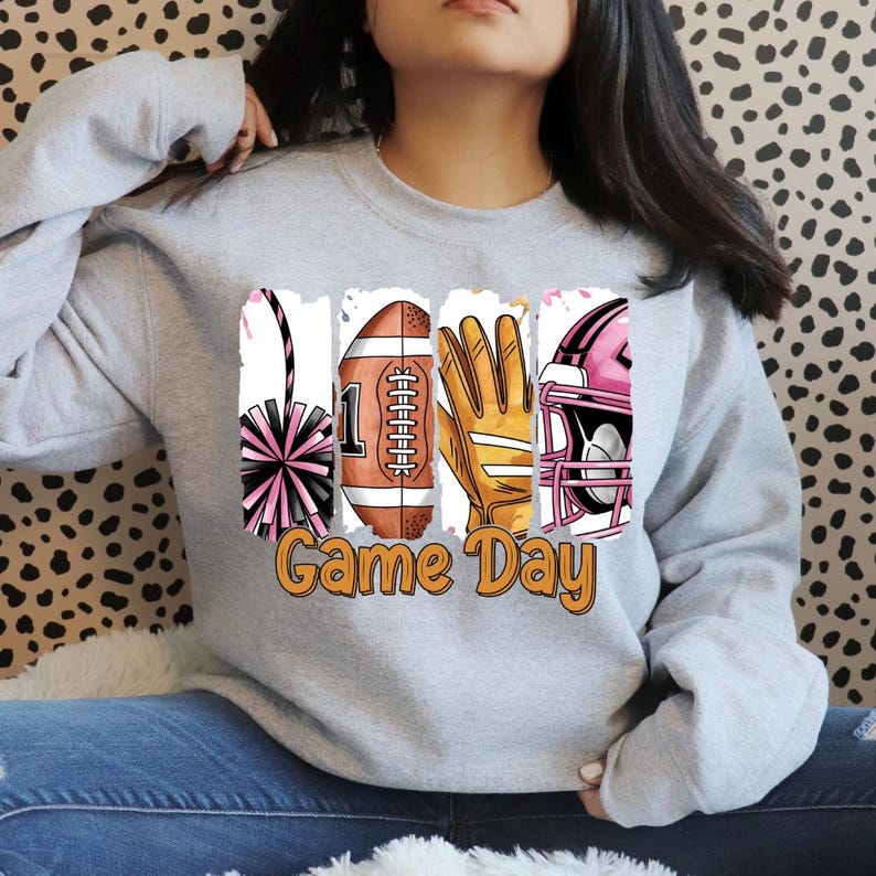 Puede incluir: Una sudadera gris claro de cuello redondo con un estampado gr&aacute;fico de cuatro paneles verticales que muestran un pomp&oacute;n rosa, un bal&oacute;n de f&uacute;tbol americano, un guante deportivo amarillo y un casco de f&uacute;tbol americano rosa, con el texto naranja Game Day.