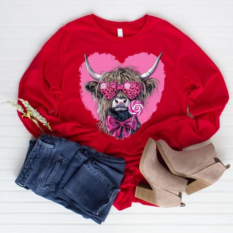 May include: A red long-sleeve shirt featuring a Highland cow wearing heart-shaped sunglasses and a pink bow tie, set against a pink heart. The cow is holding a lollipop. Blue jeans and tan boots are also in the image.